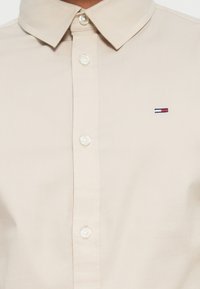 Beige skjorte med knapper og klassisk krage, med hvite knapper. Har en liten brodert logo i blått, rødt og hvitt på venstre bryst.