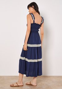 Vestido en azul marino con volantes, cuerpo fruncido, tirantes con volantes y acentos de encaje en las capas. Tela ligera, forma fluida, hasta la rodilla.
