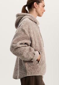 Pullover con cappuccio foderato in pile di un morbido beige, caratterizzato da una superficie testurizzata, tasche laterali e polsini a contrasto. Fit rilassato con orlo curvo.