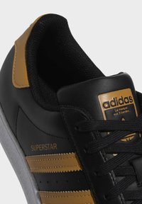 Black leather sneaker with yellow accents, featuring three stripes, textured tongue, rubber sole, and a gold logo tag. Style: Superstar.