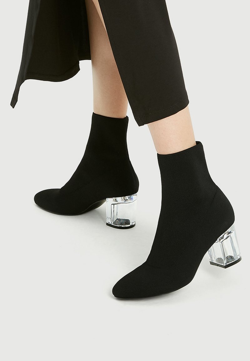 Bottines à talons hauts noires avec un design semblable à des chaussettes, dotées d'un bout pointu et d'un talon en bloc transparent. Texture lisse et silhouette ajustée.