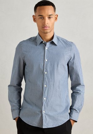 Young man wearing a blue and white vertical striped button-up shirt with long sleeves, standing against a plain light background.