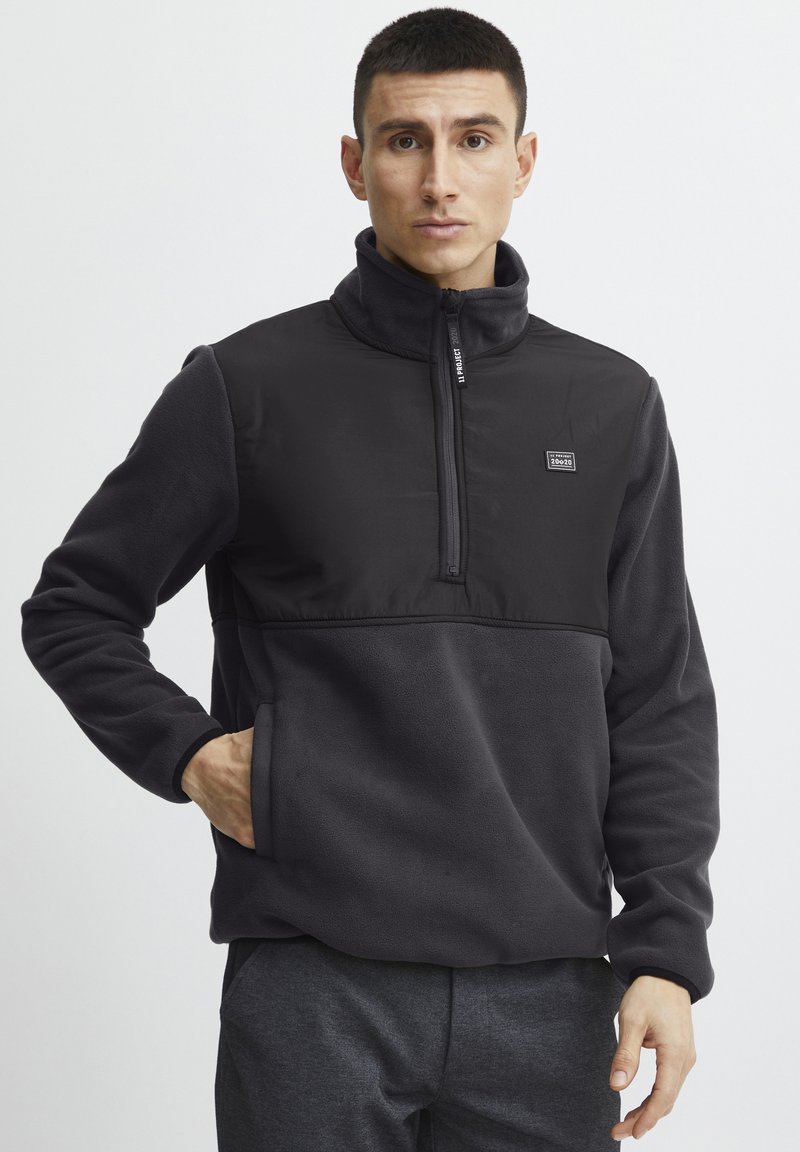 Pull en polaire noir avec zip demi-hauteur, présentant une partie supérieure lisse et une partie inférieure texturée, ainsi que des poches avant.