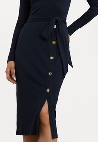 Tumsudas zilas zīmuļa svārki, kas izgatavoti no mīksta auduma, īpaši izceļ saistīto jostu, sānu šķēlumu un zelta toņa pogu apdari gar priekšpusi.