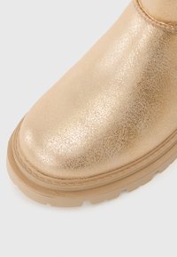 Zlaté metalické členkové topánky s textúrovaným povrchom, zaoblenou špičkou a béžovou gumovou podrážkou. Obsahujú detaily prešívania pozdĺž okraja.