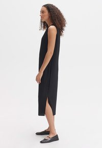 Black pleated sleeveless dress with side slits, made from a lightweight fabric. Paired with black flats, shown from the side.