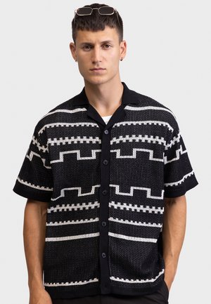 Ung mand iført sort strikket kortærmet cardigan med hvide geometriske mønstre og solbriller hvilende på hovedet mod en ensfarvet baggrund.