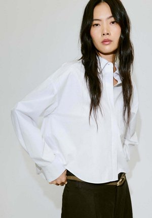 Femme aux longs cheveux noirs portant une chemise blanche boutonnée oversize et un pantalon foncé sur un fond clair uni.