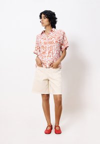 Korte mouwen, knoopshirt in het wit met een oranje abstract patroon; beige denim shorts met rafelige zoom; rode platte schoenen met uitsparingen.
