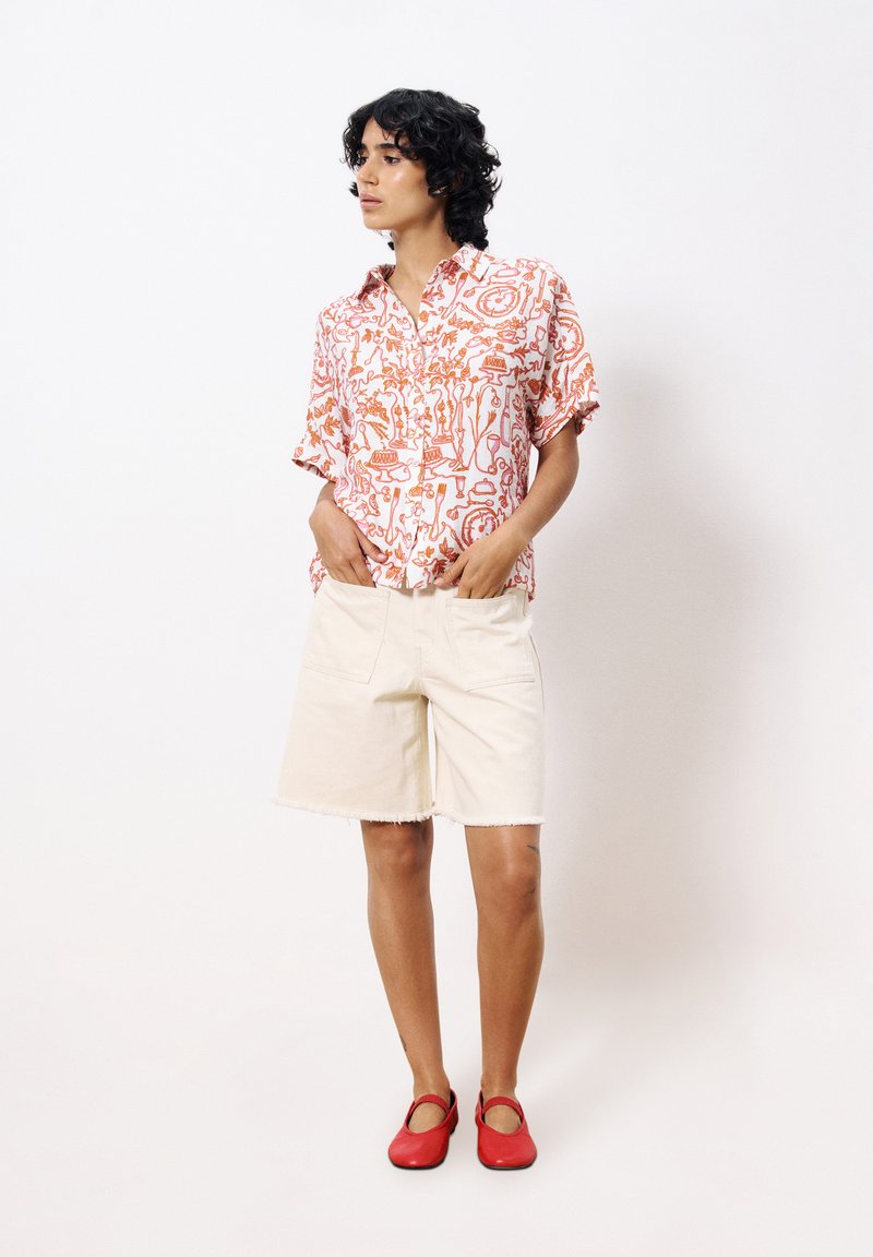 Korte mouwen, knoopshirt in het wit met een oranje abstract patroon; beige denim shorts met rafelige zoom; rode platte schoenen met uitsparingen.