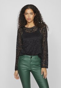 Black lace long-sleeve top with a scalloped neckline and intricate floral patterns. Paired with green faux leather high-waisted pants.