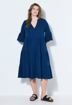 Kobieta stoi na białym tle, ubrana w luźną, granatową sukienkę do kolan z rozkloszowanymi rękawami oraz czarne sandały z odkrytymi palcami.