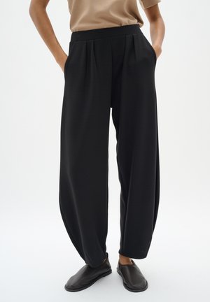 Pantalons noirs amples avec plis, en tissu texturé, dotés de poches latérales et d'une ceinture large. Associés à des chaussures noires à enfiler.