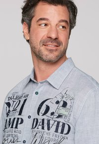 Camisa abotonada en tejido de rayas azul y blanco. Presenta impresiones de logotipo y texto en azul oscuro, botones en contraste y un cuello extendido.