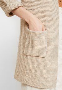 Hand placed inside the front pocket of a beige knitted cardigan with rolled-up sleeves and a textured stitch pattern.