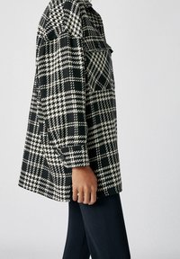 Kurtka w czarno-białą kratę o luźnym kroju, z kołnierzem, zapinanymi mankietami oraz dwiema kieszeniami bocznymi.
