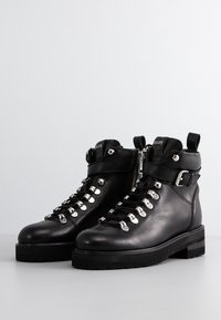 Bottines en cuir noir avec œillets argentés, bride à boucle, fermeture éclair latérale et semelles à crampons épaisses sur un fond neutre.