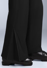 Černé voděodolné kalhoty s rozšířeným střihom, vybavené zipovými detaily na lemech a hladkým povrchem, kombinované s černými botami.