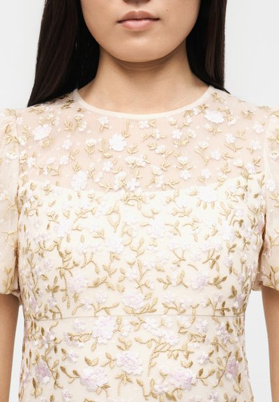 Γυναίκα που φοράει ελαφρύ μπεζ τοπ με κοντά μανίκια, διακοσμημένο με κεντητά λευκά και χρυσά ανθικά σχέδια πάνω σε ύφασμα.
