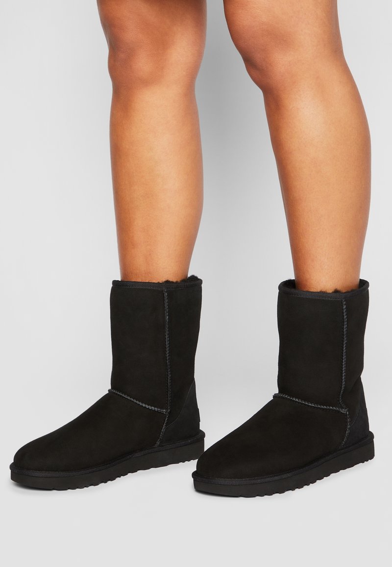 Personne portant des bottes mi-mollet en daim noir avec des détails de couture, debout sur une surface gris clair.