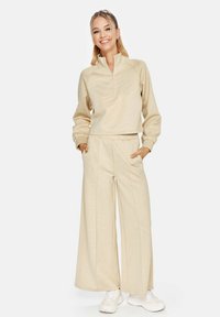 Beige zip-up sweatshirt met ribgebreide manchetten, gecombineerd met wijde, bijpassende broeken. Zachte textuur en subtiele patroon details, gedragen met witte sneakers.