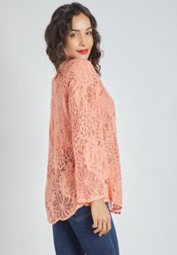 Kimono de encaje coral con patrón floral, mangas largas y dobladillo de puntilla. Combinado con jeans azules, que destacan su textura transparente.