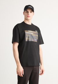 Svart t-skjorte med korte ermer og grafisk design som har gule prikker og abstrakte linjer, sammen med en svart caps som viser hvit tekst.