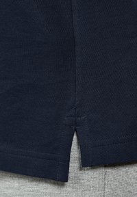 Granatowa bawełniana koszulka polo z teksturowanym wykończeniem, z bocznymi rozporkami i prostym dołem. Szary materiał widoczny pod koszulką.