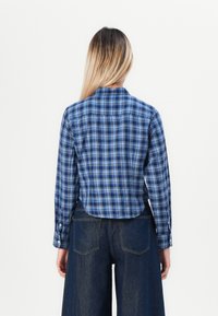 Blå rutete skjorte med knapper og lange ermer, kort design, med hvite knapper og ryggdetalj, sammen med mørke vide bukser.