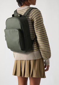 Un sac à dos vert en matériau lisse. Dispose de poches zippées, de bretelles rembourrées et d'une poignée robuste. Porté avec un pull rayé et une jupe plissée.