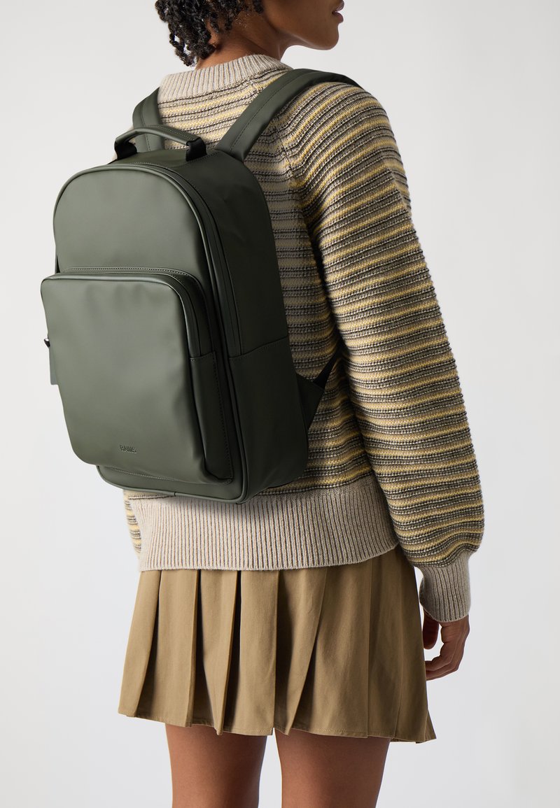 Un sac à dos vert en matériau lisse. Dispose de poches zippées, de bretelles rembourrées et d'une poignée robuste. Porté avec un pull rayé et une jupe plissée.