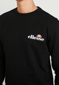 Sudadera de algodón negra con un cuello redondo, que presenta un logo "ellesse" en blanco y un gráfico colorido de medio sol bordado en el pecho.