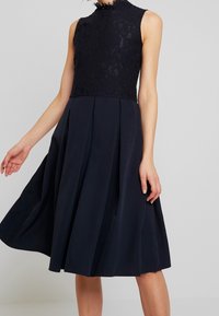 Marinblå klänning med en spetsbodice och hög halsringning. Plisserad kjol ger volym, mjuk textur i tyget, ser knälång ut.