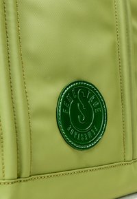 Borsa in tessuto verde con design strutturato, logo rotondo prominente in verde scuro e dettagli in cucitura beige. Texture liscia.