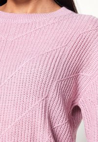 Ljusrosa stickad tröja med ribbat mönster, rund halslinning och diagonala sömmar som löper över tyget.