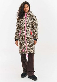 Pružný kabát s leopardím potiskem a kapucí, růžový zip, kapsy a šňůrky, v kombinaci s hnědými kalhotami a černými nazouvacími botami.