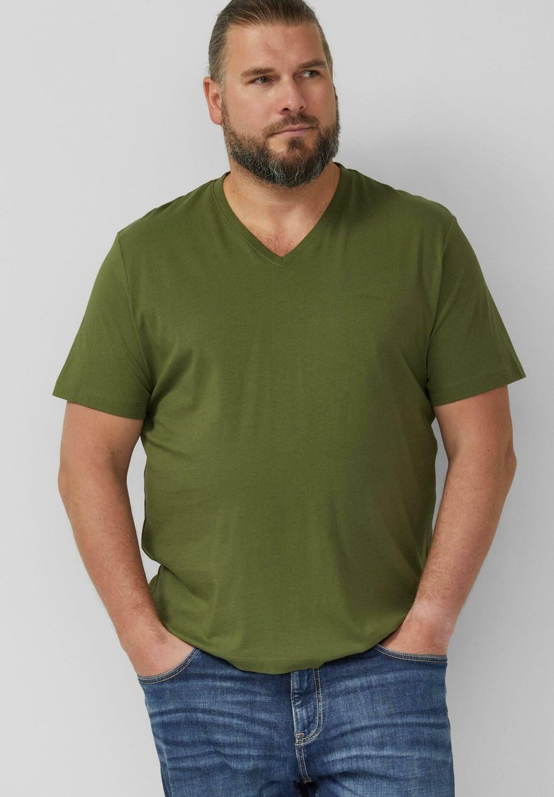 Homme barbu portant un t-shirt à col en V vert olive et un jean bleu, debout les mains dans les poches devant un fond uni.