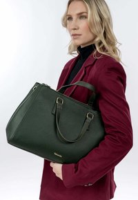Grøn tekstureret læder tote taske med korte håndtag og hardware i guldtoner, båret af en person iført en bordeaux blazer og sort top.
