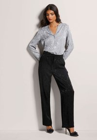 Zilveren, gestructureerde blouse met een V-hals en lange mouwen, gecombineerd met zwarte, loszittende, patroongedrukte broek en zwarte hoge hakken.