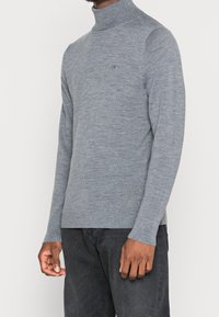 Pull col roulé gris en tissu doux et texturé, avec des manches longues et un logo sur la poitrine. Porté avec un jean noir.