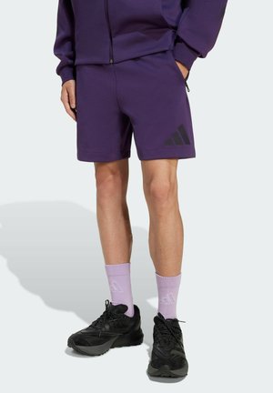 Paarse sportieve shorts van zacht materiaal met een zwart logo aan de zijkant; gecombineerd met lichtpaarse sokken en zwarte sneakers.