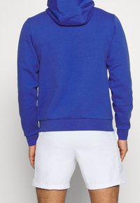 Felpa blu con una vestibilità morbida, caratterizzata da un cappuccio con cordino e polsini a coste, abbinata a pantaloni corti bianchi. Tessuto dalla consistenza liscia.
