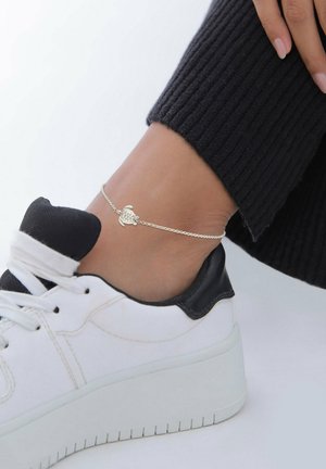 Ankel med sølvskildpadde-charmearmbånd båret over en hvid sneaker med sort kant og sorte ribstrikkede bukser.