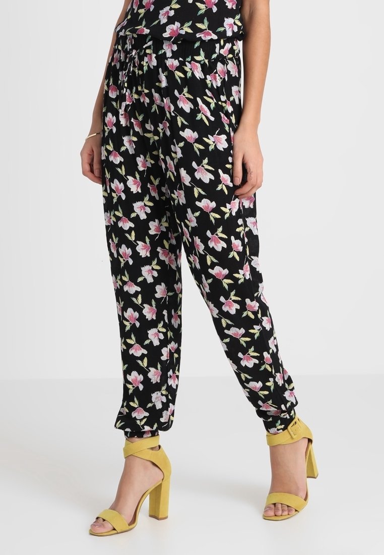 Femme portant un pantalon noir à motif floral et des sandales jaunes à talons hauts, debout devant un fond clair uni.