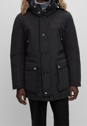 Czarna ocieplana zimowa parka z zapięciem na guziki i zamek błyskawiczny, cztery kieszenie z klapkami oraz kaptur obszyty futrem, noszona na szarym swetrze z golfem.