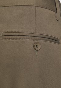 I pantaloni elasticizzati marroni presentano una tasca posteriore singola con chiusura a bottone. Il tessuto ha una texture liscia e un design elegante.