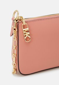 Pieni vaaleanpunainen nahkakotelo kultaisella vetoketjulla, kultaisella MK-logolla vetoketjun vetimessä ja kultaisella ketjukiinnityksellä valkoista taustaa vasten.