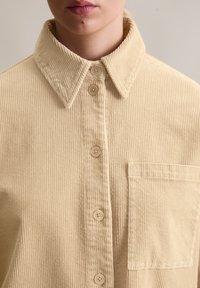 Beżowa koszula ze sztruksu z ostrym kołnierzykiem, guzikami z przodu i jedną kieszenią na piersi. Teksturowana tkanina z pionowymi prążkami.