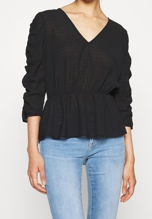 Femme portant un chemisier texturé noir à col en V avec des manches longues froncées et un jean bleu clair sur un fond uni.