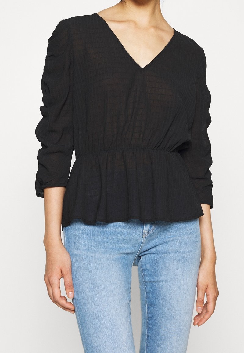 Femme portant un chemisier texturé noir à col en V avec des manches longues froncées et un jean bleu clair sur un fond uni.